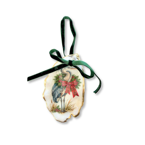 Decoupage oyster shell ornament with a blue heron wearing an evergreen wreath and plaid bow, gilded rim, and dark green velvet ribbon by Melissa Fouts.