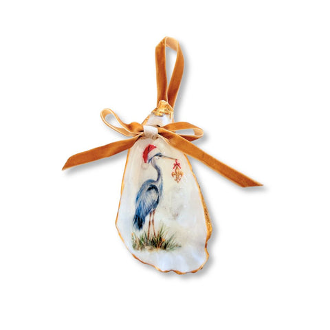 Decoupage oyster shell ornament with a blue heron in a Santa hat holding a fleur-de-lis, gilded rim, and gold velvet ribbon by Melissa Fouts.